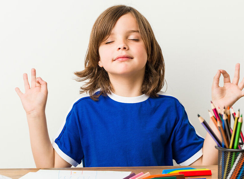 Kind sitzt mit geschlossenen Augen am Tisch und meditiert, daneben liegen bunte Stifte und Papier