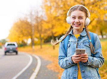 Ein lächelndes Mädchen mit Kopfhörern geht im Herbst an einer Strasse entlang. Sie hat ein Smartphone in der Hand.