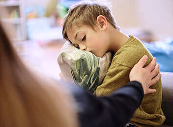 Ein Junge sitzt traurig auf einem Sofa und hält ein Kissen im Arm. Eine erwachsene Person legt ihm tröstend die Hand auf die Schulter.