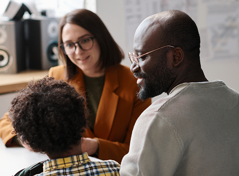 Ein Mann sitzt mit seinem Kind bei einem Gespräch mit einer Frau in einem Büro. Die Frau lächelt, während sie mit den beiden spricht. Im Hintergrund sind Regale, Lautsprecher und Unterlagen zu sehen.