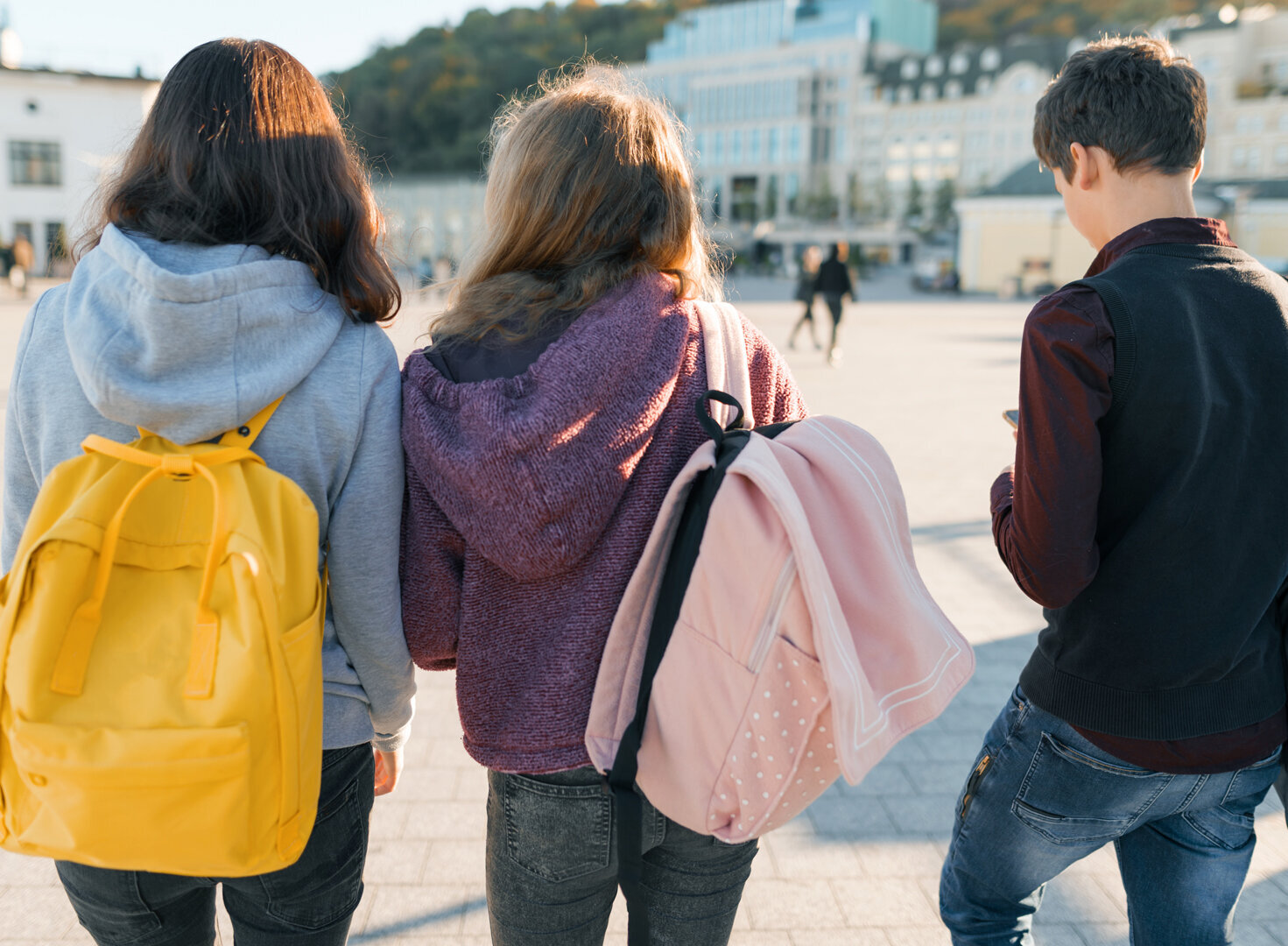 3 Teenager laufen über einen Platz. Man sieht sie von hintern. Sie tragen Rucksäcke.