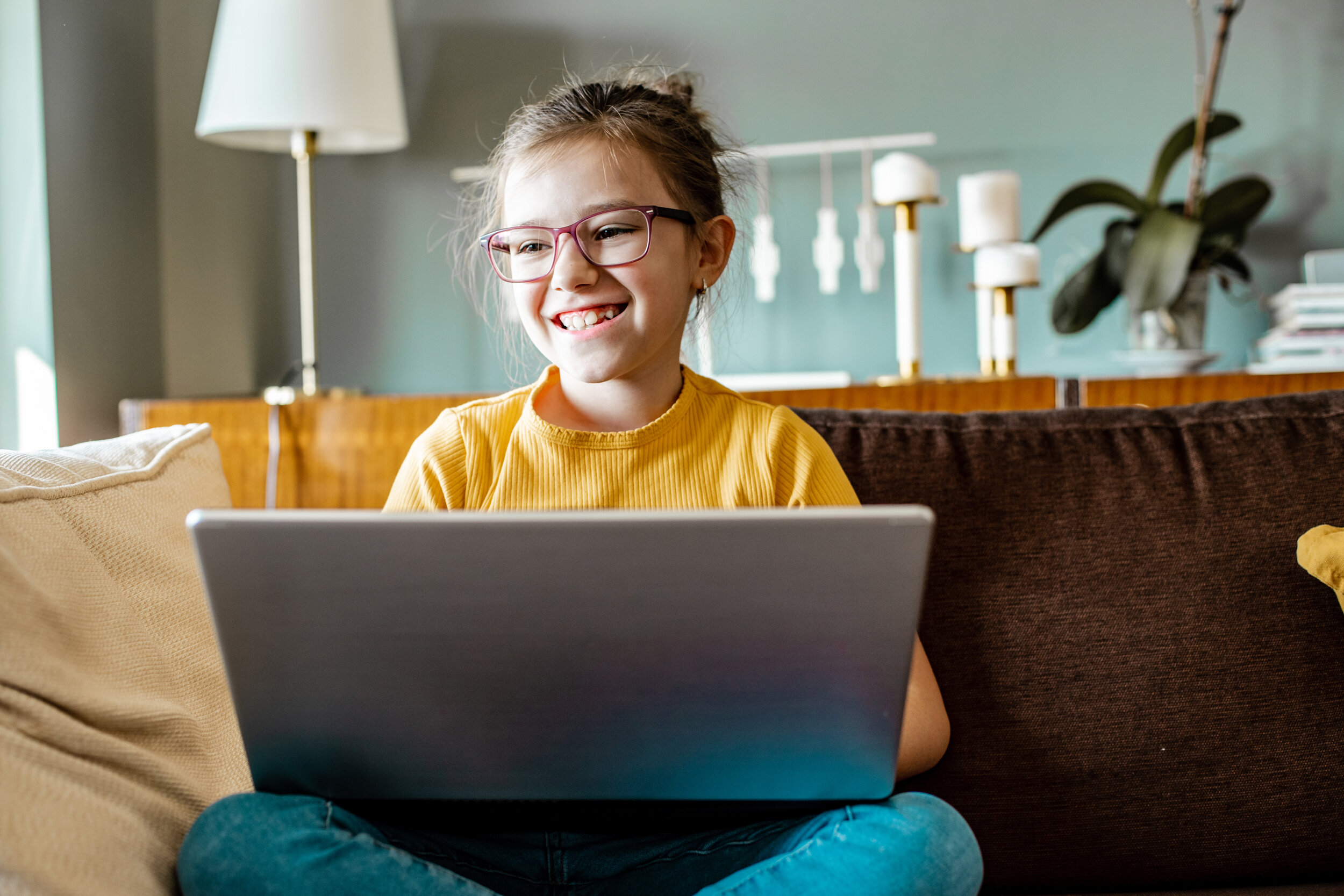 Schülerin mit rosafarbener Brille arbeitet konzentriert und lächelnd am Laptop auf Sofa