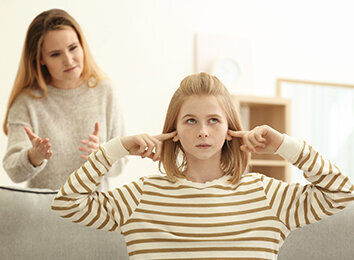 Mädchen hält sich die Ohren zu. Hinter ihr schimpft eine Frau mit ihr.