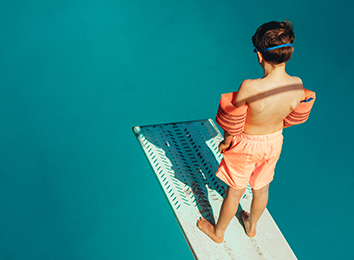Junge mit Schwimmflügeln steht auf Sprungbrett im Schwimmbad und schaut ins Becken.