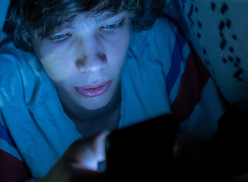 Ein Junge schaut unter der Bettdecke auf sein Handy.