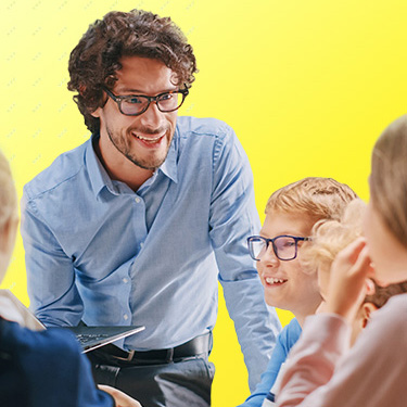 Lehrer in blauem Hemd spricht lächelnd mit einer Gruppe von Schülerinnen und Schülern an einem Tisch.