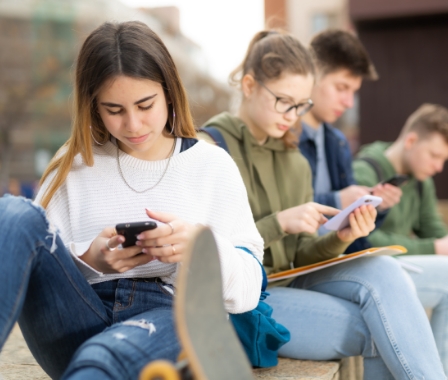 Vier Jugendliche sitzen im Freien, alle schauen auf Smartphones oder Tablets. Im Vordergrund ein Mädchen in weißem Pullover mit Skateboard.