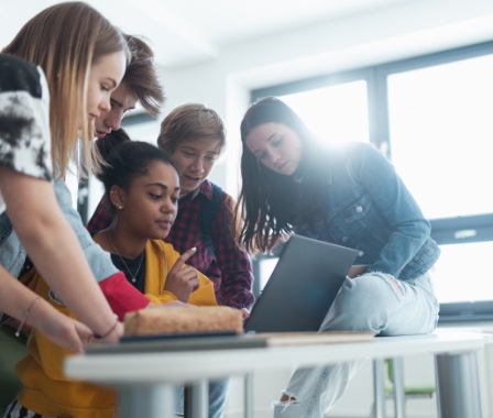 Fünf Jugendliche stehen und sitzen um einen Tisch und schauen gemeinsam auf den Bildschirm eines Laptops.