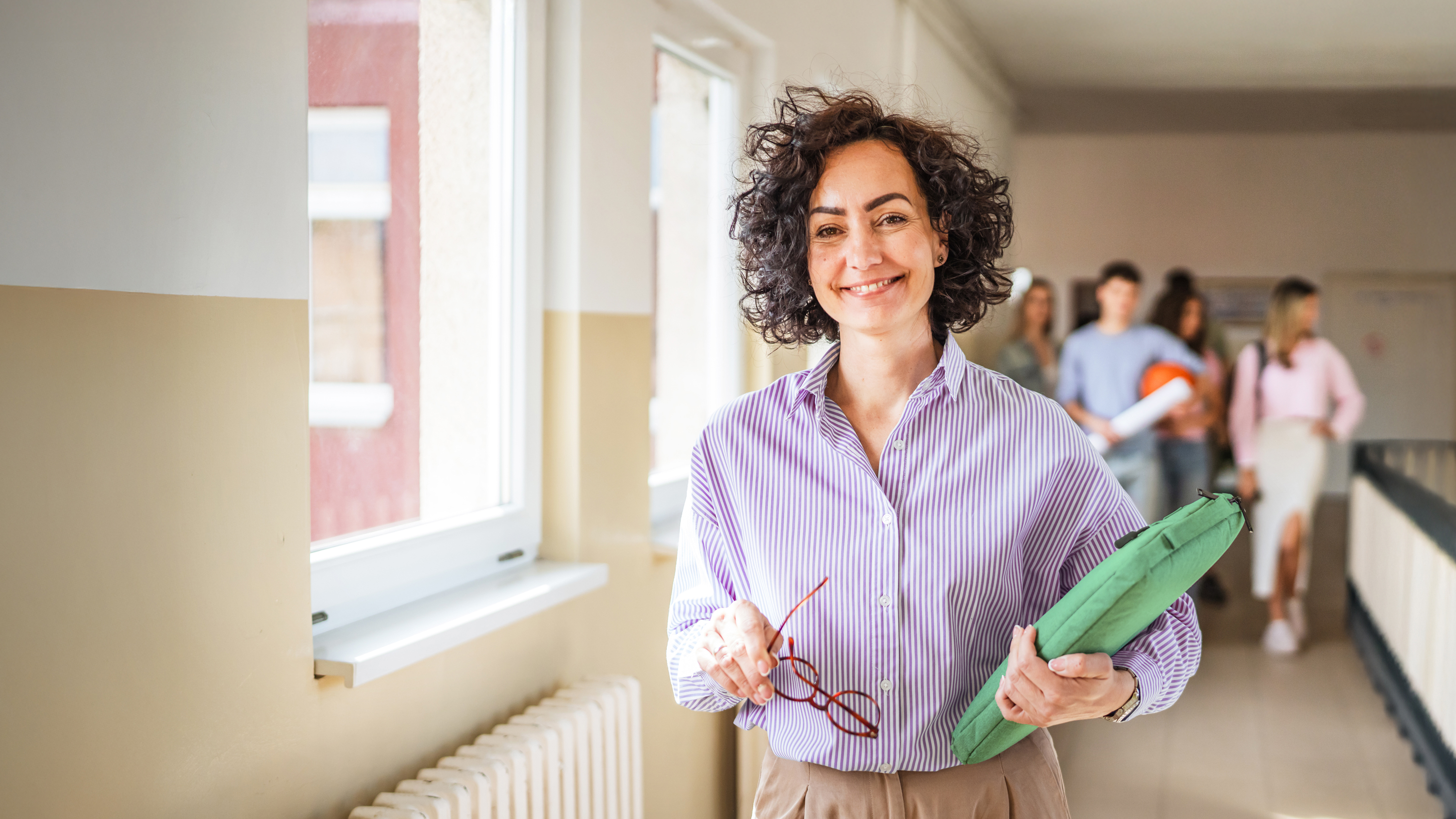 Lehrerin mit grünem Ordner und Brille im Schulflur mit Schüler:innen, moderne Schulumgebung, digitale Medienkompetenz